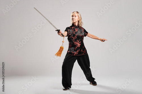 Mature blonde woman practicing Tai Chin with sword in a white background wearing a traditional chinese black jacket with red chinese decoration, black trousers and chinese shoes with ying yang symbol