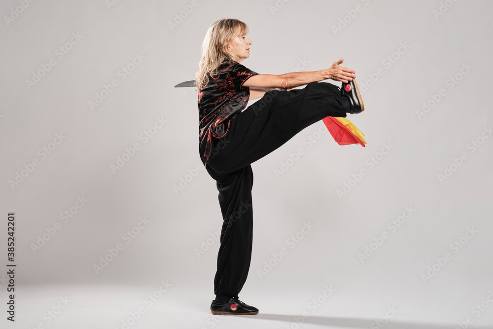 Mature blonde woman practicing Tai Chin with saber in a white background wearing a traditional chinese black jacket with red chinese decoration, black trousers and chinese shoes with ying yang symbol