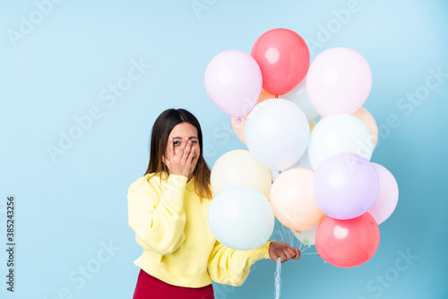 Woman holding balloons in a party over isolated blue background covering eyes and looking through fingers