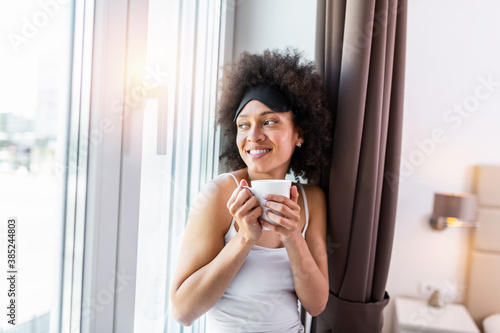 Young and cute lady sitting on the windowsill and looking out the window with cup of coffee in the morning. Young woman drinking coffee and wearing sleeping mask