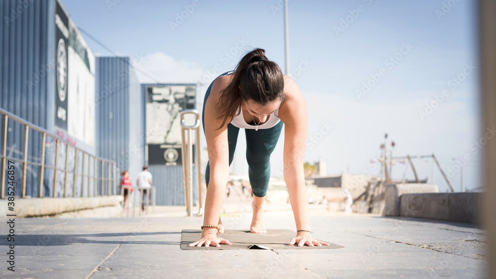 Fototapeta premium Sporty young woman doing Yoga exercises using a gym mat along the beach in Lisbon, Portugal. Playful woman working as freelance Yoga teacher doing fitness workout on the beach at sunset. Healthy life