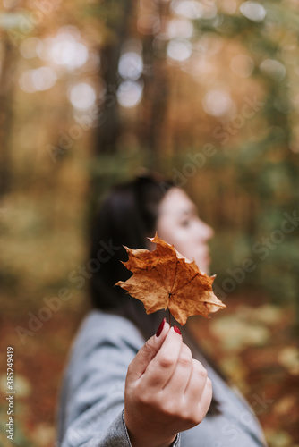 A beautiful woman in a gray coat in autumn with a maple leaf in her hand. Blurry portrait of a woman