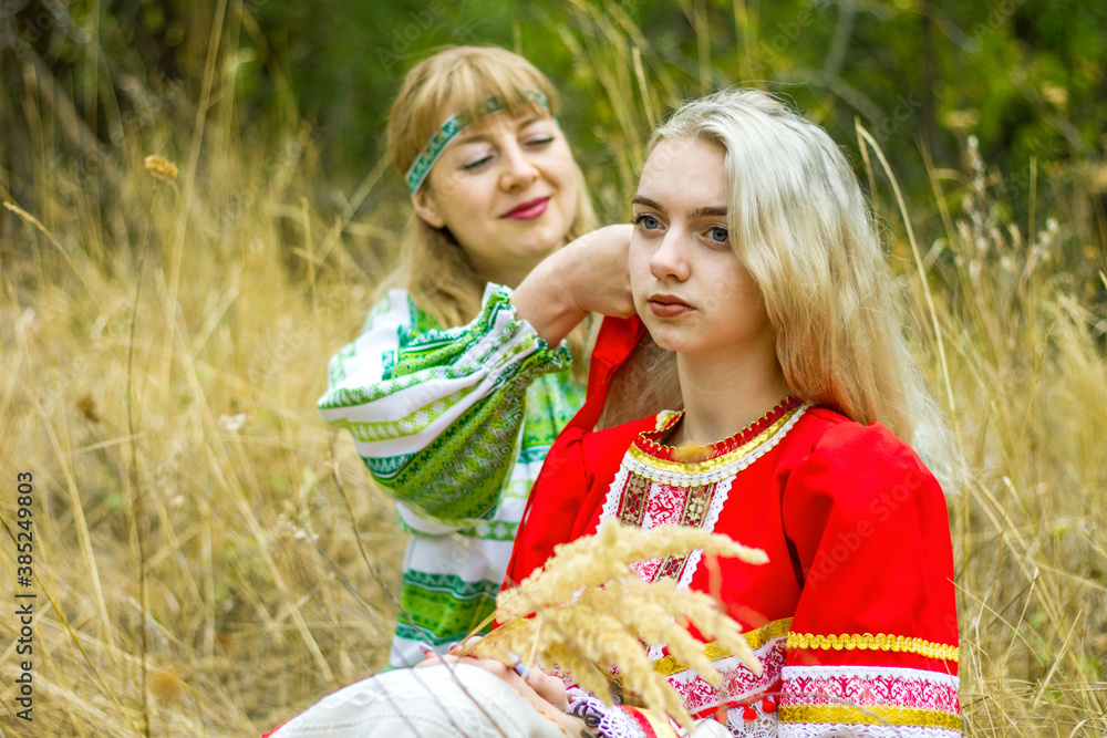 A young woman in a green folk costume is braiding a braid for a blonde girl in a red folk costume, the girls are sitting in the grass in the forest.