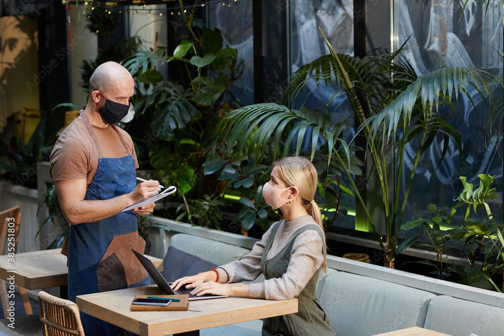 Businesswoman in protective mask working at the table with laptop and making an order to the waiter in mask theey are in the restaurant Businesswoman in protective mask working at the table with laptop and making an order to the waiter in mask theey are in the restaurant