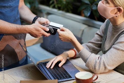 Young businesswoman in protective mask paying with credit card while sitting at the table in front of laptop in cafe