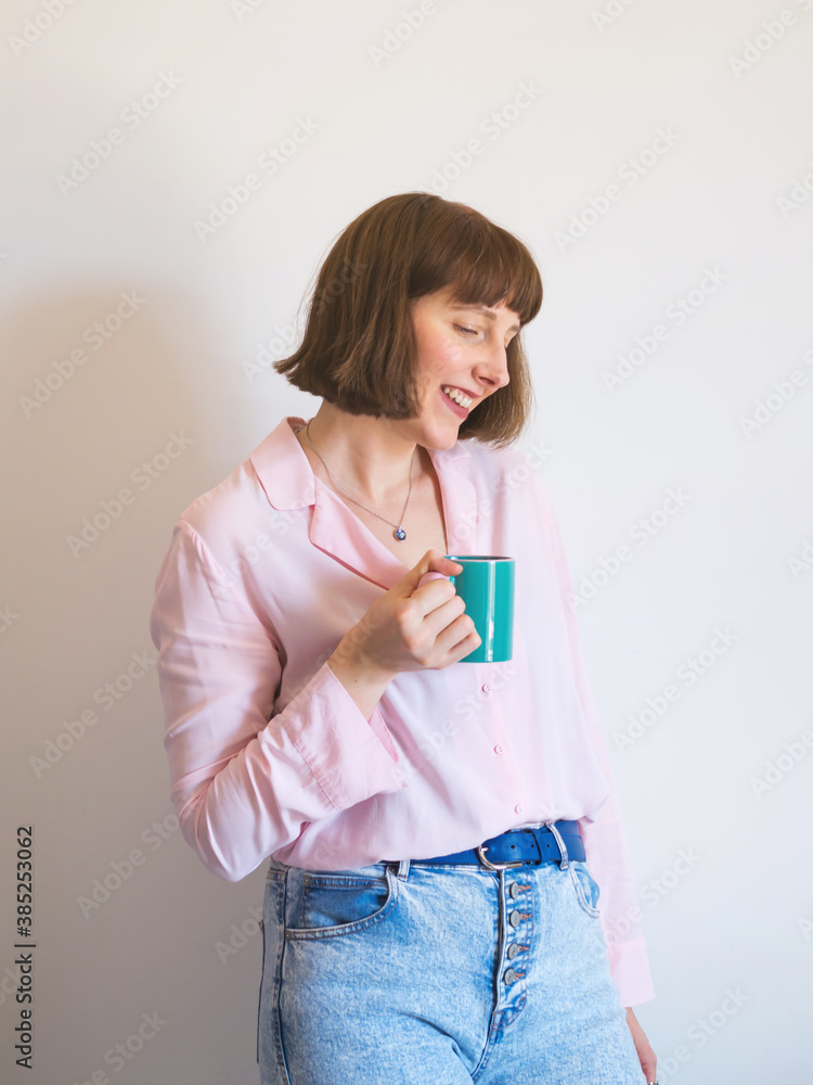 Young woman wearing jeans and pink shirt drinking coffee and smiling over white wall background. Young woman wearing jeans and pink shirt drinking coffee and smiling over white wall background.