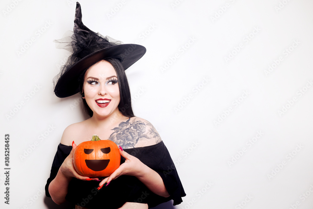 A girl dressed as a witch is holding a pumpkin. Model in hat with tattoo on shoulder smiling A girl dressed as a witch is holding a pumpkin. Model in hat with tattoo on shoulder smiling