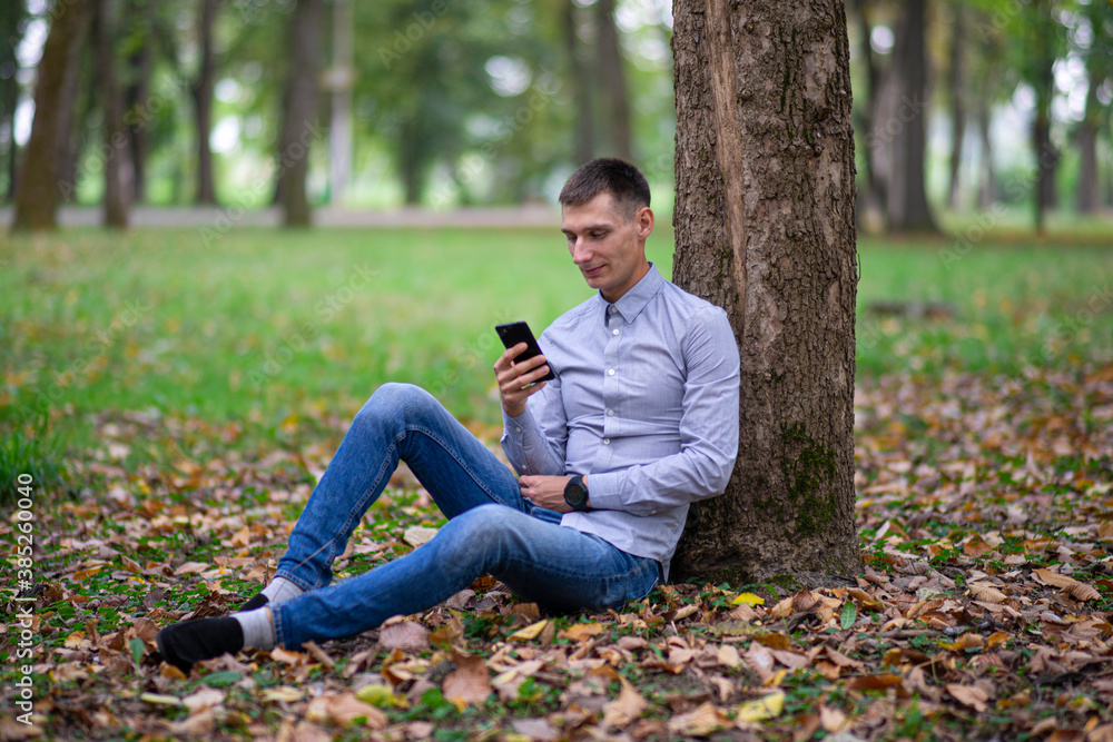 Portrait of a young man in an autumn park, near a tree, outdoors on a natural green background. Serious guy looks at the phone, smiling. Communication concept. Selective focus