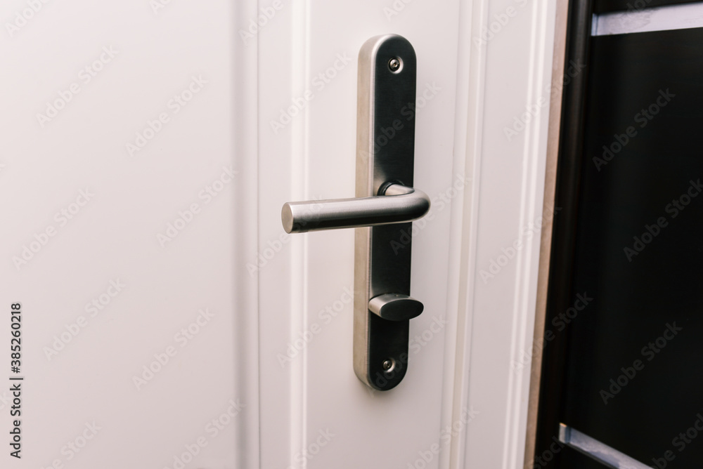 White door and handle. Modern chrome handle in your hotel room or home. Entrance to an apartment, office, or bedroom. Detail of a wooden door. Interior of a house or hotel White door and handle. Modern chrome handle in your hotel room or home. Entrance to an apartment, office, or bedroom. Detail of a wooden door. Interior of a house or hotel