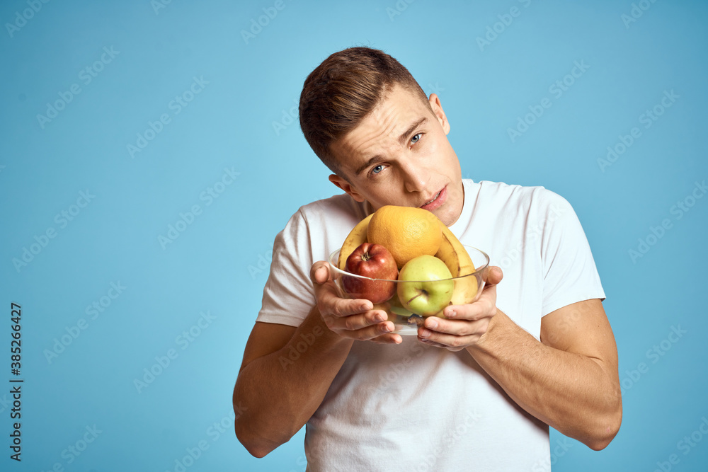Happy man with fresh fruits gesturing with hands blue background white t-shirt vitamins bananas oranges apples Happy man with fresh fruits gesturing with hands blue background white t-shirt vitamins bananas oranges apples