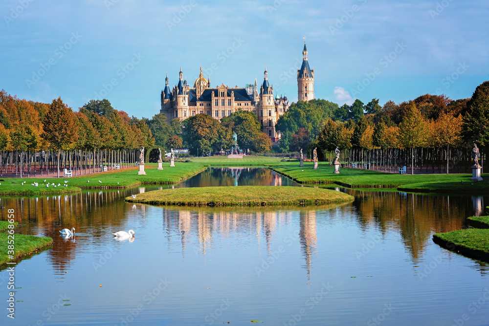 Schweriner castle seen from the park with water canals, reflections, sculptures and trees with colored foliage on a sunny autumn day, copy space