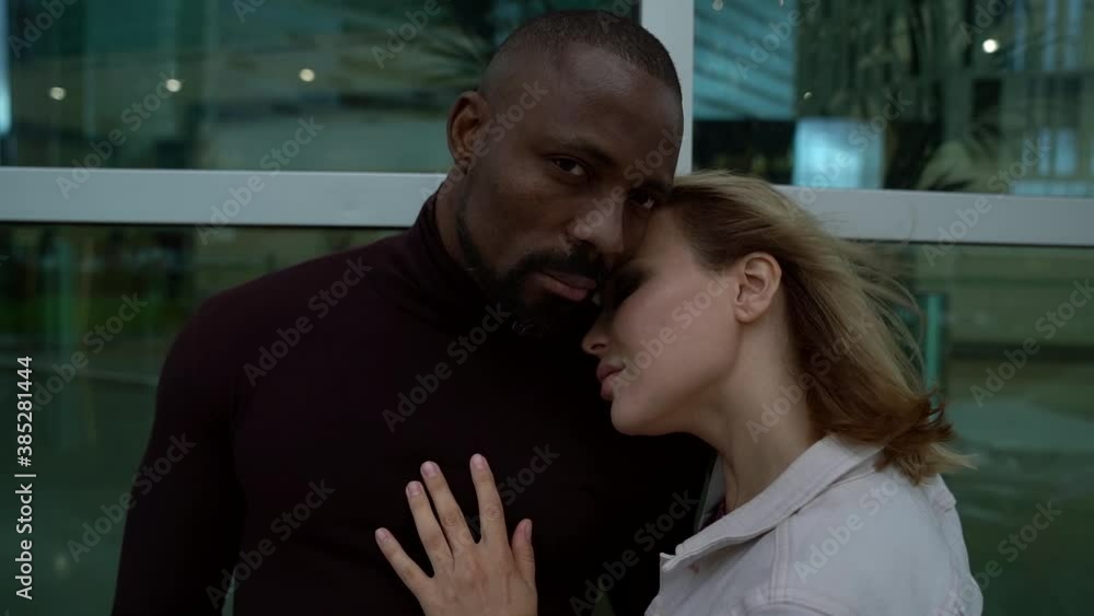an African man in dark clothing and a blonde woman in a gray jacket are hugging each other and standing against a glass wall. the woman put her head on the guy's shoulder an African man in dark clothing and a blonde woman in a gray jacket are hugging each other and standing against a glass wall. the woman put her head on the guy's shoulder