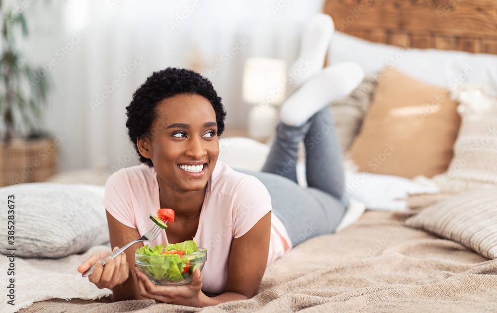 Healthy vegan diet. Smiling black lady lying on bed at home with bowl of fresh vegetable salad, copy space. Panorama Healthy vegan diet. Smiling black lady lying on bed at home with bowl of fresh vegetable salad, copy space. Panorama