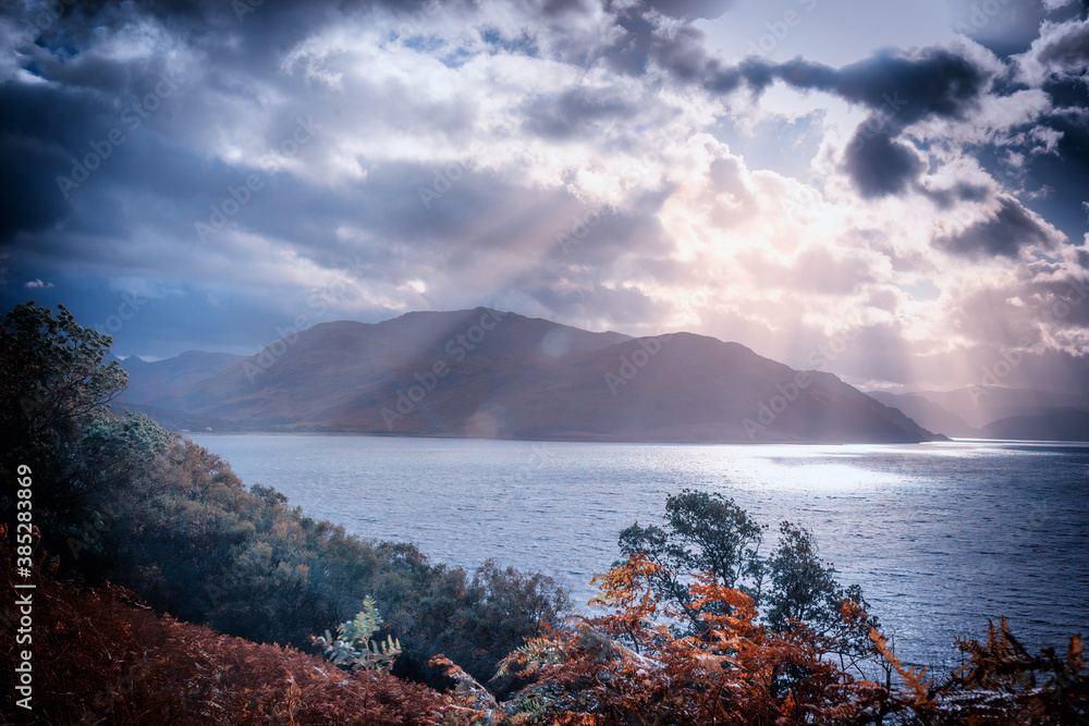 Rays of sunlight break through brooding storm clouds to illuminate a Scottish Loch and the trees and bracken in the foreground the mountains in the background loom in the shadows cast by the clouds