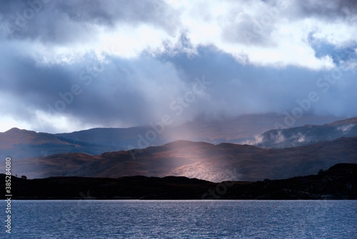 A ray of sunlight breaks through brooding storm clouds to illuminate a small patch of a Scottish loch shore the trees glowing in the golden light contrasted against the dark shoreline and Loch water
