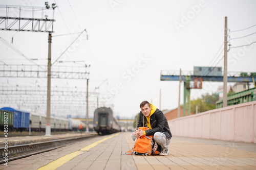Young man squats on platform, waiting for train. Male passenger with backpacks sitting on railroad platform in waiting for train ride. Concept of tourism, travel and recreation.
