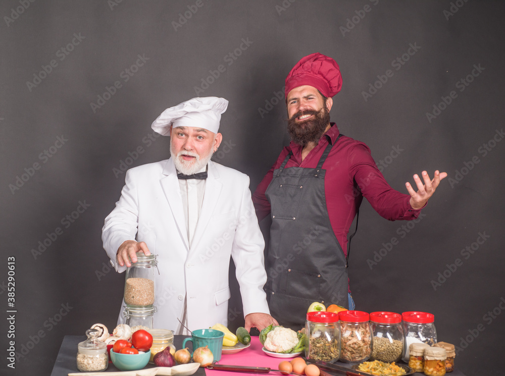 Kitchen. Cooking. Two chefs on kitchen. Beared chef man. Delicious food. Male chef in uniform. Satisfied bearded chef, cook or baker. Kitchen. Cooking. Two chefs on kitchen. Beared chef man. Delicious food. Male chef in uniform. Satisfied bearded chef, cook or baker.