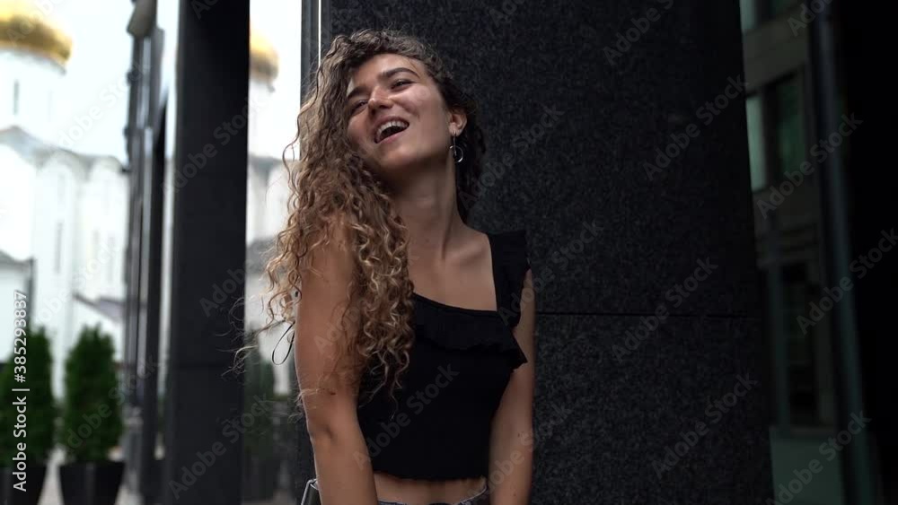 a young woman with curly hair leans against a dark gray wall and laughs. bottom view. camera zooms in a young woman with curly hair leans against a dark gray wall and laughs. bottom view. camera zooms in