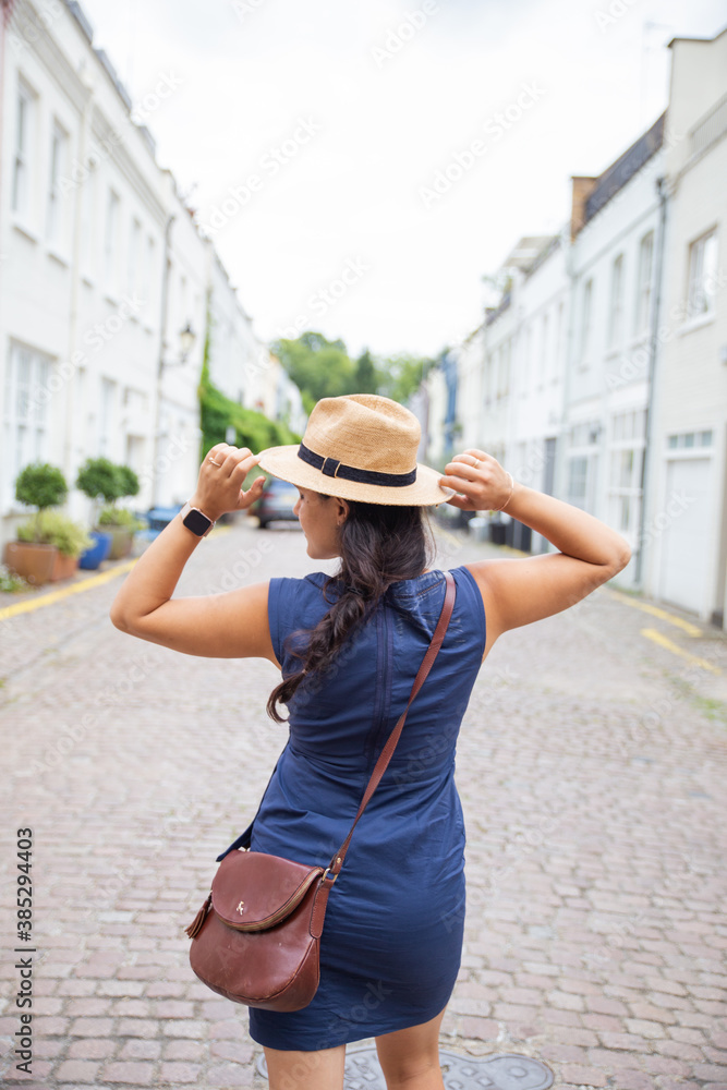 Smiling Woman in Blue Dress From Behind, Calmly Adjusts Her Hat with Both Hands