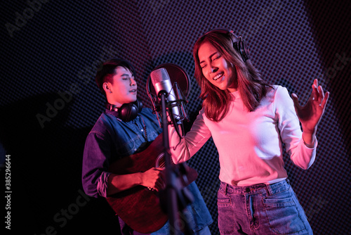 Pretty Asian female singer recording songs by using a studio microphone and pop shield on mic with male playing guitar in blue and red light. Performance and show in the music business. Duet session.