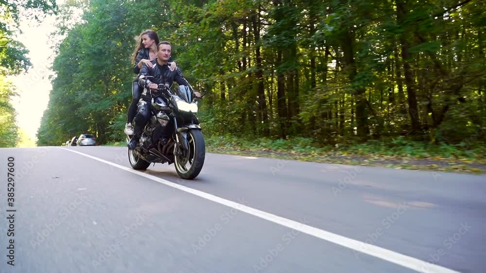 young happy couple in love motorcyclists in black leather jackets on a sports bike ride on the road smiling and hugging. Young sexy woman with boyfriend drive. Lifestyle. through the woods and trees young happy couple in love motorcyclists in black leather jackets on a sports bike ride on the road smiling and hugging. Young sexy woman with boyfriend drive. Lifestyle. through the woods and trees