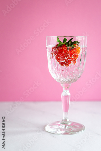 A single glass with a strawberry with bubbles on pink background and marble surface, vertical