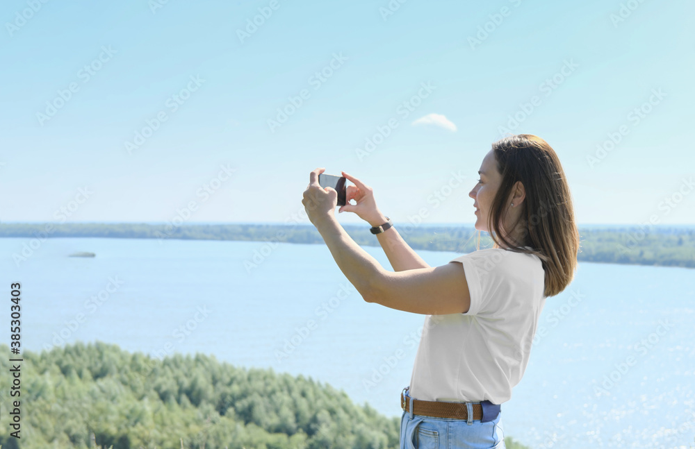 beautiful woman blogger taking a photo on a smartphone. happy woman over river landscape, travelling and sightseeing. local travel. weekend vacation. casual outfit, jeans and t-shirt. beautiful woman blogger taking a photo on a smartphone. happy woman over river landscape, travelling and sightseeing. local travel. weekend vacation. casual outfit, jeans and t-shirt.
