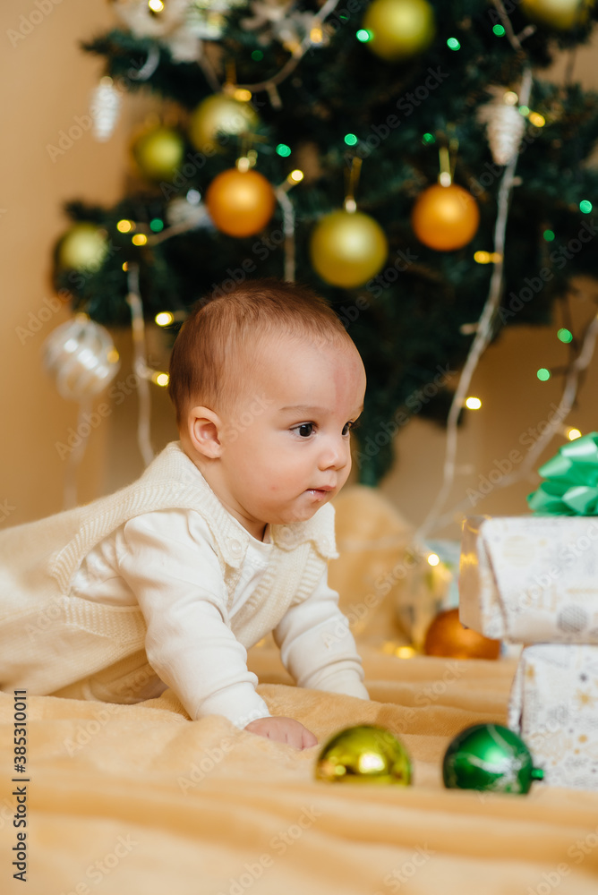 Obraz premium Cute smiling baby is lying under a festive Christmas tree and playing with gifts. Christmas and New Year celebrations