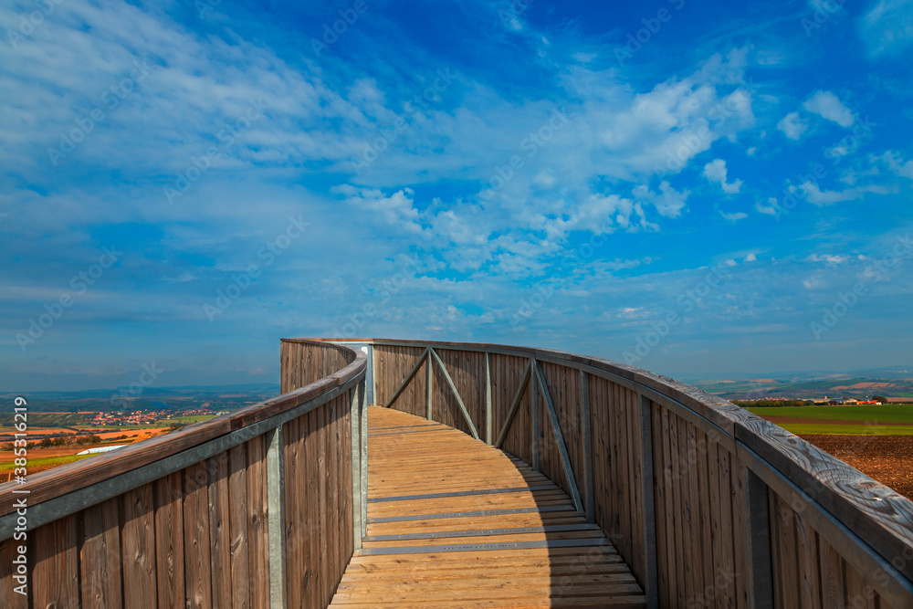 Fototapeta premium A round footbridge to the lookout tower in Kobyla in Moravia in the Czech Republic. There is a view of the countryside from the lookout tower. In the background is a blue sky with white clouds.