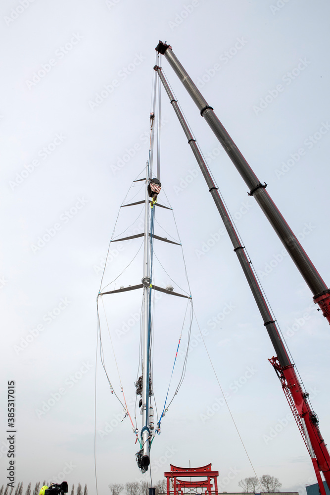 Placing a mast at a super sailing yacht at the harbor. Hoisting. Lifting a mast of a ship with a crane. Shipbuilding industry. Placing a mast at a super sailing yacht at the harbor. Hoisting. Lifting a mast of a ship with a crane. Shipbuilding industry.