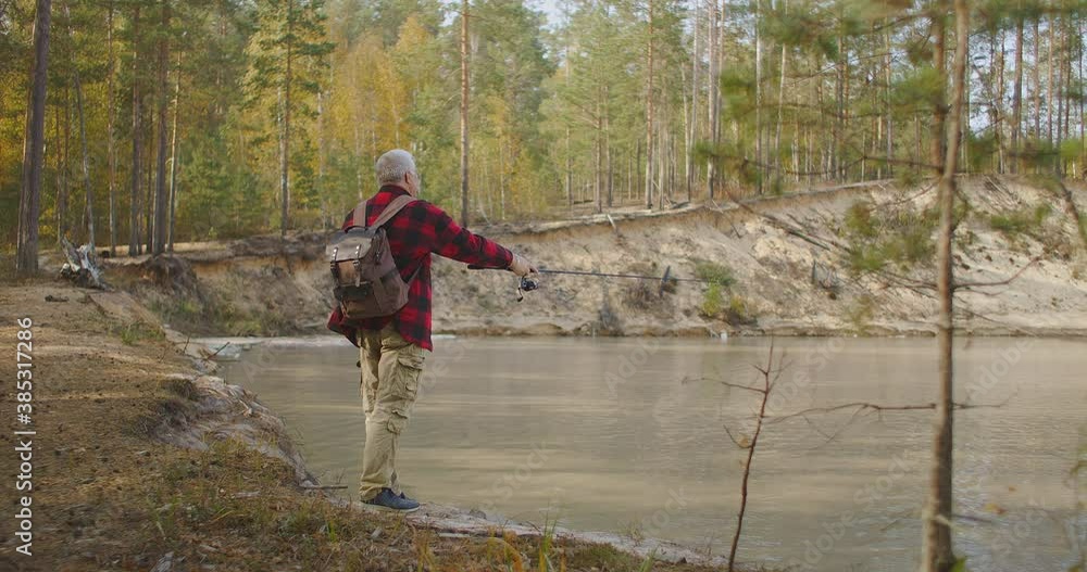 grey-haired man dressed red checkered shirt is casting fishing rod in river, relaxing on shore of reservoir