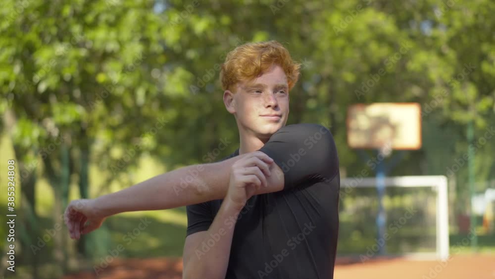 Confident young sportsman warming up before training outdoors and leaving. Middle shot portrait of smiling Caucasian redhead man stretching neck and hands on sunny outdoor court and walking away. Confident young sportsman warming up before training outdoors and leaving. Middle shot portrait of smiling Caucasian redhead man stretching neck and hands on sunny outdoor court and walking away.