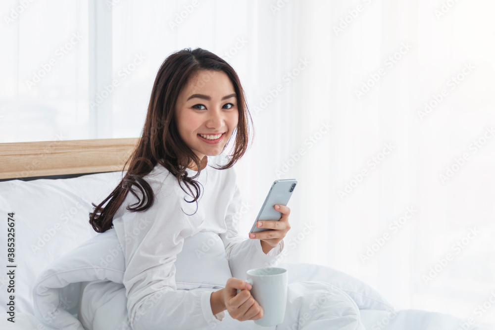 Young woman in white relaxing and drinking cup of hot coffee or tea using smartphone on bed in the bedroom. Happy girl using a mobile phone on bed. Beautiful woman using smartphone in the morning. Young woman in white relaxing and drinking cup of hot coffee or tea using smartphone on bed in the bedroom. Happy girl using a mobile phone on bed. Beautiful woman using smartphone in the morning.