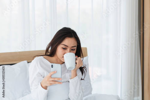 Young woman in white relaxing and drinking cup of hot coffee or tea using smartphone on bed in the bedroom. Happy girl using a mobile phone on bed. Beautiful woman using smartphone in the morning.