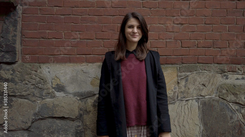 Portrait of cute young shy woman standing against red brick wall looking around and smiling for camera