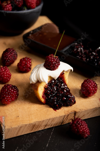 Muffin de mora cubierto de crema chantilly partido por la mitad sobre tabla y fondo negro al lado de cobertura de chocolate.