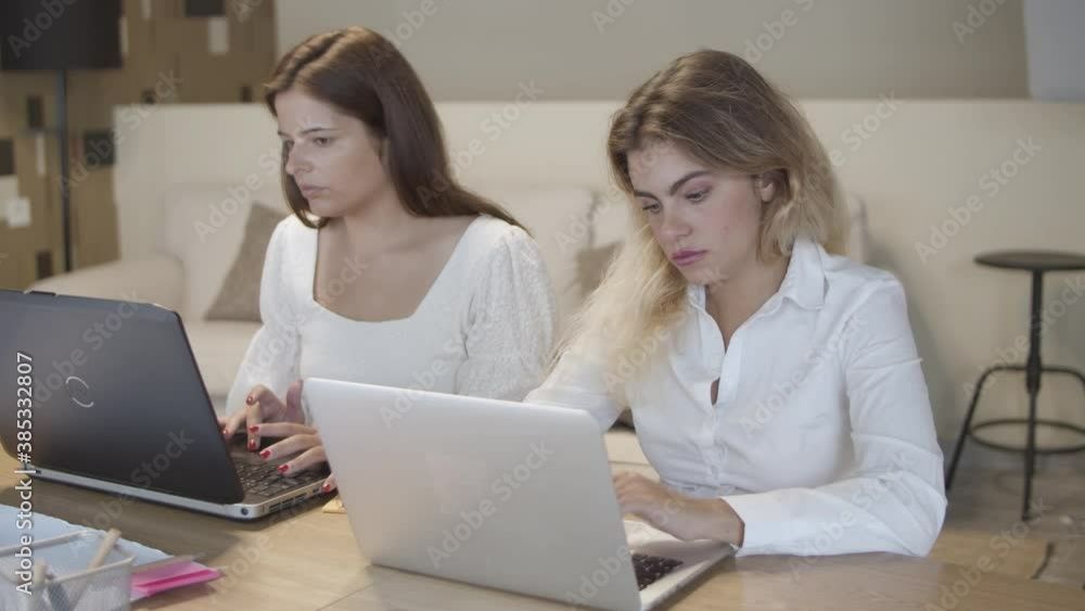 Two female managers sitting together at table, working in silence, typing on laptops, chatting online. Medium shot. Co-working and communication concept Two female managers sitting together at table, working in silence, typing on laptops, chatting online. Medium shot. Co-working and communication concept