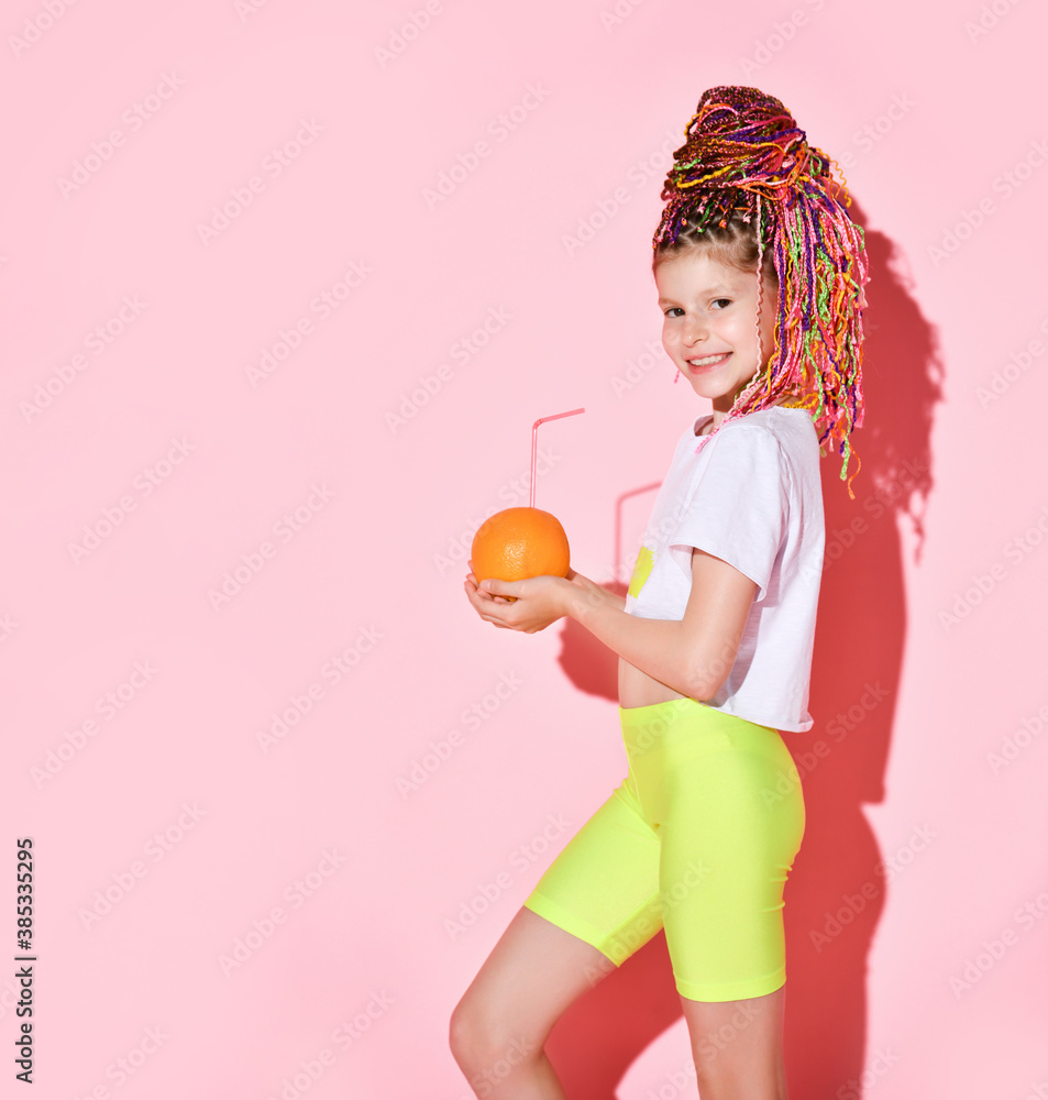 Teenage girl with african pigtails is holding orange and drinking it through tubule, posing sideways on pink background. Close up