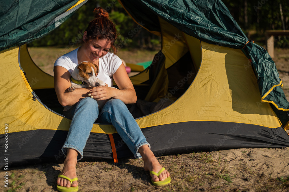 Beautiful red-haired woman is resting in nature with her pet. A girl sits in a tourist tent and hugs a Jack Russell Terrier in the forest. The dog and its female owner at the campsite.
