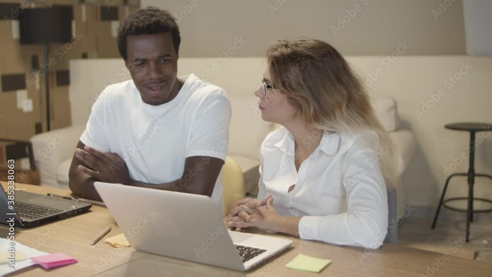 Diverse couple of colleagues sitting at table with laptops and talking, discussing project in co-working space. Medium shot. Corporate communication concept Diverse couple of colleagues sitting at table with laptops and talking, discussing project in co-working space. Medium shot. Corporate communication concept