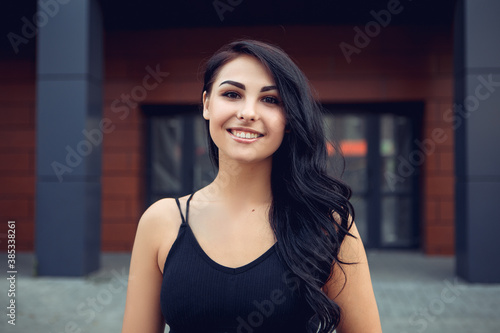 Portrait of young smiling positive woman standing outside, dressed in black singlet having wide white smile, looking at the camera. Image with copy space.
