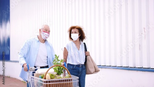 Senior couple with face masks and shopping walking outside supermarket in city, coronavirus concept. photo