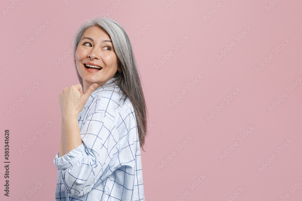 Side view of cheerful funny gray-haired asian woman wearing basic white checkered shirt standing pointing thumb aside on mock up copy space isolated on pastel pink colour background, studio portrait. Side view of cheerful funny gray-haired asian woman wearing basic white checkered shirt standing pointing thumb aside on mock up copy space isolated on pastel pink colour background, studio portrait.