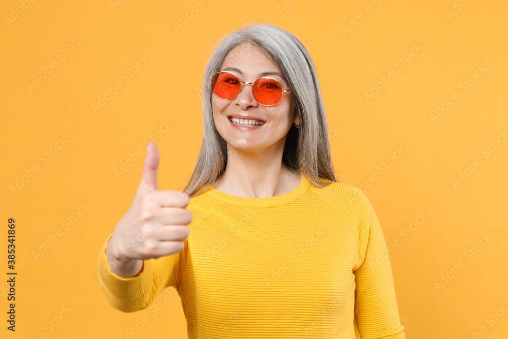 Smiling cheerful beautiful attractive gray-haired asian woman wearing casual clothes eyeglasses standing showing thumb up looking camera isolated on bright yellow colour background, studio portrait. Smiling cheerful beautiful attractive gray-haired asian woman wearing casual clothes eyeglasses standing showing thumb up looking camera isolated on bright yellow colour background, studio portrait.