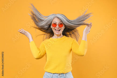 Excited cheerful cute gray-haired asian woman in casual clothes eyeglasses spreading hands throwing flowing hair having fun looking camera isolated on bright yellow colour background studio portrait.