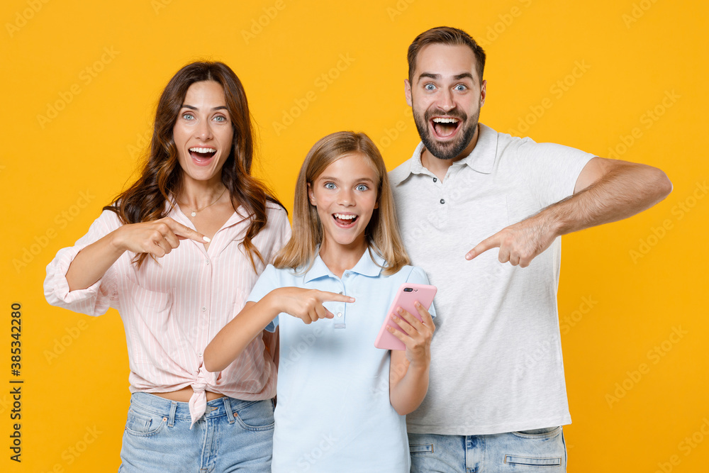 Obraz premium Surprised funny young parents mom dad with child kid daughter teen girl in basic t-shirts pointing index fingers on mobile cell phone isolated on yellow background studio portrait. Family day concept.