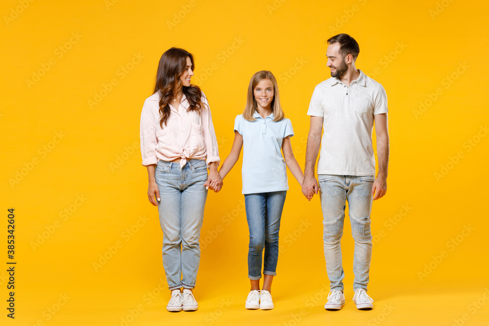 Full length portrait of smiling young parents mom dad with child kid daughter teen girl in t-shirts holding hands looking at each other isolated on yellow background. Family day parenthood concept. Full length portrait of smiling young parents mom dad with child kid daughter teen girl in t-shirts holding hands looking at each other isolated on yellow background. Family day parenthood concept.