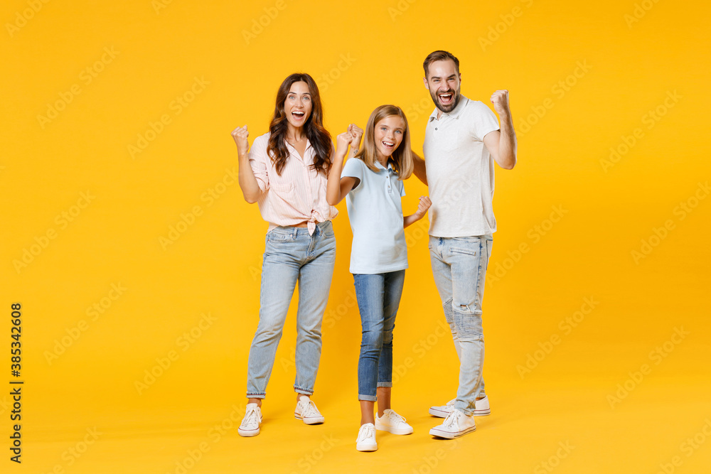 Obraz premium Full length portrait happy joyful laughing young parents mom dad with child kid daughter teen girl in t-shirts clenching fists doing winner gesture isolated on yellow background. Family day concept.