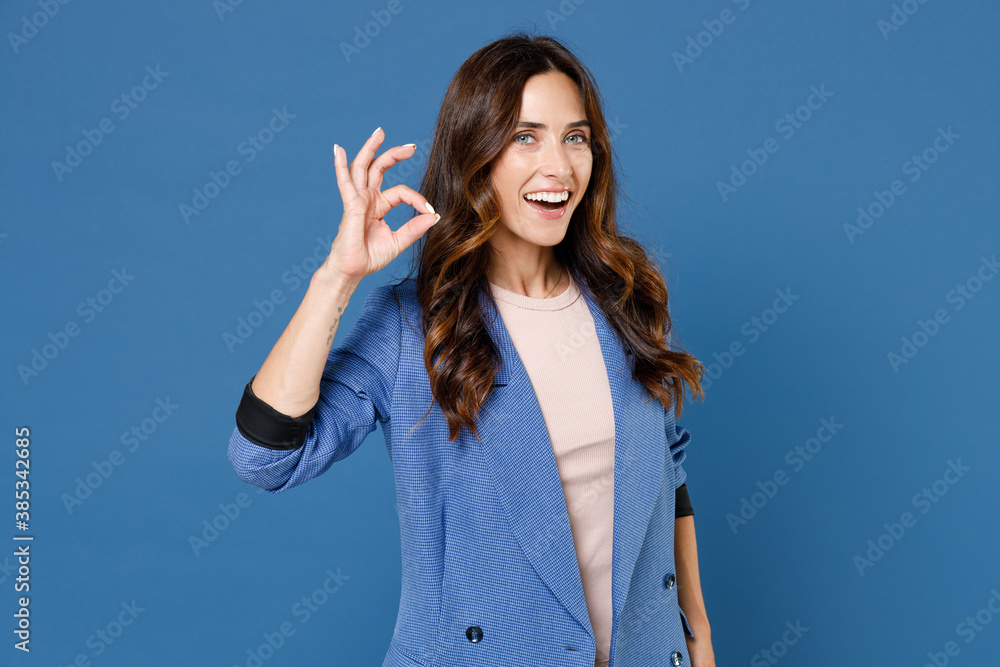 Excited cheerful laughing funny pretty attractive young brunette woman 20s wearing basic jacket standing showing OK gesture looking camera isolated on bright blue colour background, studio portrait. Excited cheerful laughing funny pretty attractive young brunette woman 20s wearing basic jacket standing showing OK gesture looking camera isolated on bright blue colour background, studio portrait.