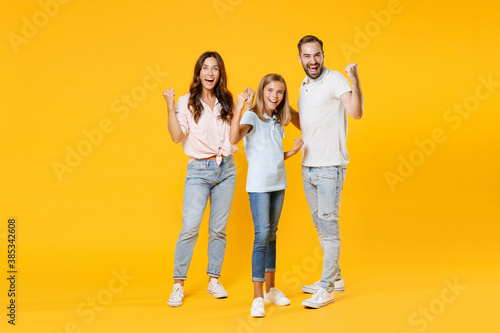 Full length portrait happy joyful laughing young parents mom dad with child kid daughter teen girl in t-shirts clenching fists doing winner gesture isolated on yellow background. Family day concept.
